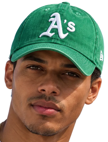 Two models wearing green Oakland Athletics baseball caps with white 'A's' lettering and a white New Era logo, set against a clear blue sky.