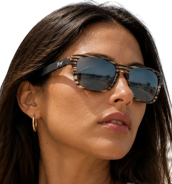 a woman wearing tortoiseshell sunglasses with dark lenses, photographed outdoors against a blue sky background.