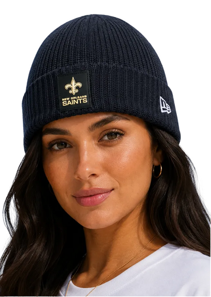 A woman wearing a dark navy ribbed knit beanie with a New Orleans Saints patch on the front and a white New Era logo on the side, set against a solid light blue background.